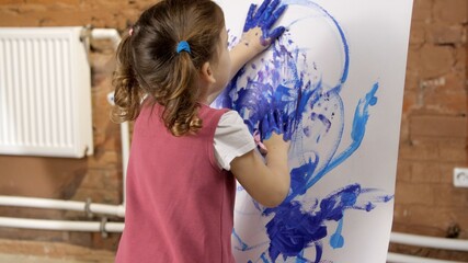 Pretty little girl draw with palms in blue paint on canvas on an easel. Painted hands leave a mark on the paper. Children's creativity. Close-up shot of painting process. Child draws with fingers. 