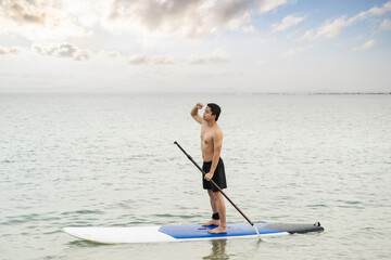 Fototapeta na wymiar Asian man is paddling on a SUP board, standup paddler at the ocean during sunset, summer holidays vacation travel.