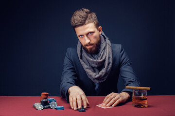 Stylish bearded Man in suit and scarf playing in dark casino, smoking cigar, drink whiskey.