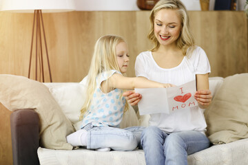 Happy mother day in sunny flat. Child daughter congratulates mom and gives her postcard with heart drawing. Family concept