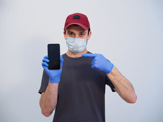 Fototapeta na wymiar A courier service man in a medical mask and blue protective gloves is holding a phone isolated on a white background. Phone blank screen with place for text, announcements, discounts. Online shopping
