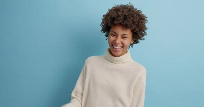 Pretty overjoyed dark skinned woman laughs out as hears something hilarious feels very happy and upbeat communicates with best friend expresses positive emotions isolated over blue background