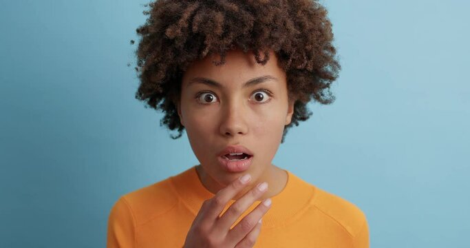 Stunned dark skinned woman looks impressed at camera stares bugged eyes and opens mouth with amazement being speechless dressed casually isolated over blue background. Human emotions concept