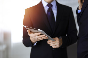 Business people use a tablet computer for discussion of their new project, standing in sunny modern office. Unknown businessman or male entrepreneur with a colleague at workplace