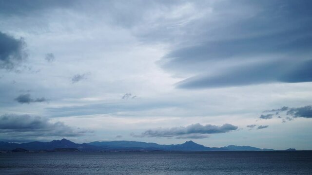 Time lapse of clouds moving over calm sea water, overcast day. Hilly coastline on horizon.