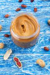 Peanut butter in an open jar and peanuts in the skin are scattered on the blue table. Space for...