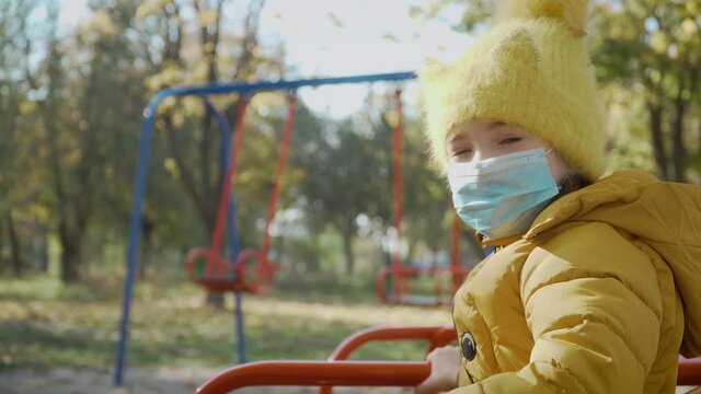 Portrait of little girl in medical mask looks sadly in child playground during worldwide virus epidemic. Stop COVID-19 infection