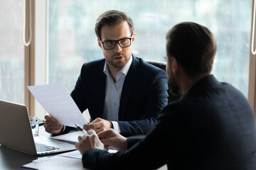 Serious businessmen sit at desk in office talk consider financial paperwork at meeting together. Concentrated male business partners brainstorm discuss company project or idea. Teamwork concept.