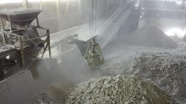 production workshop, crushing stones, huge bucket pick up stone blocks and lifts  into powerful unit. fractions and sorting on carpet line. rays of sun through dusty workshop, chemical industry grind
