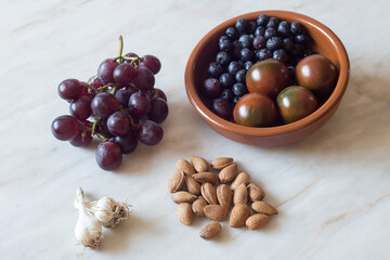 A clay bowl with blueberries and tomatoes from the garden along with a bunch of black grapes, a few heads of garlic, and a handful of shelled almonds. Various fruits and vegetables on a marble table. 