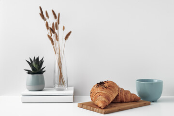 Croissant and a mug on a wooden stand, a vase and a pot with a plant on the books, white background. Eco-friendly materials in interior decor, minimalism. Copy space, mock up