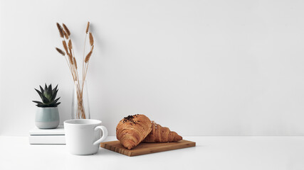 Croissant and a mug on a wooden stand, a vase and a pot with a plant on the books, white background. Eco-friendly materials in interior decor, minimalism. Copy space, mock up