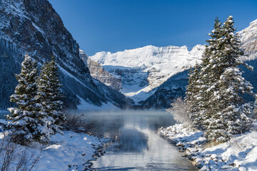 Fototapeta na wymiar Lake Louise in early winter sunny day morning. Mist floating on turquoise color water surface. Clear blue sky, snow capped mountains in background. Beautiful natural landscape in Banff National Park.