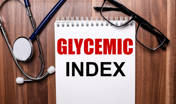 GLYCEMIC INDEX is written on white paper on a wooden background near a stethoscope and black-framed glasses. Medical concept
