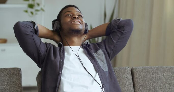 Portrait of calm young african businessman ethnic student wear wireless headphone relax with eyes closed meditate sitting sofa with folded hands behind head listen lounge music audio book course mp3