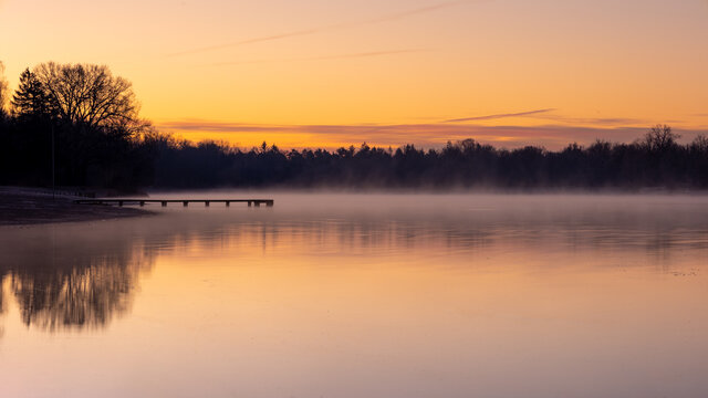 Seenlandschaft am fr&uuml;hen Morgen. Leichter Nebel habt sich stimmungsvoll vom rot der aufgehenden Sonne ab.