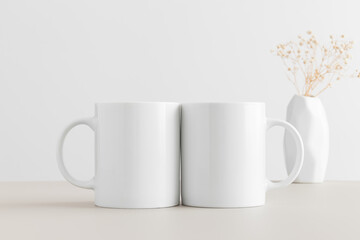 Two white mugs mockup with a gypsophila in a vase on a beige table.