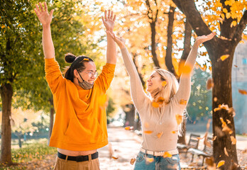 Happy smiling couple with face mask down, throwing autumn fall orange red leaves in park forest outdoor in coronavirus time - New normal seasonal activity lifestyle outside