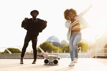 Young Afro friends dancing outdoor while listening to music with wireless headphones and vintage...