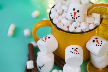 marshmallow snowmen on a mint turquoise background with a yellow cocoa mug illuminated by a garland