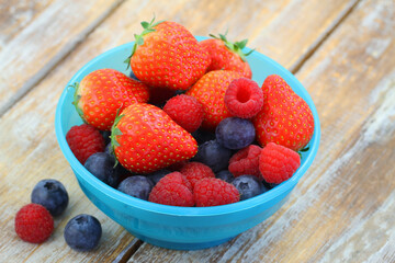 Best source of Vitamin C: strawberries, bluberries and raspberries in blue plastic bowl on wooden surface

