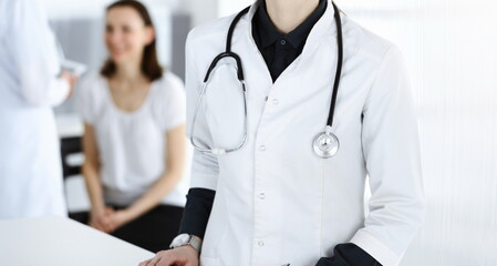 Woman-doctor at work with colleague and patient at background. Middle aged female physician filling up medical documents or prescription while standing in hospital reception desk. Data in medicine