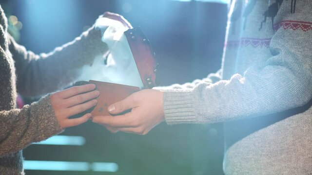 Man presenting a xmas gift to a woman. Close up of hands present a cascet with faity light and smoke on garland bokeh background. Merry christmas and happy new year concept. Celebrating holidays