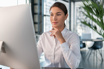 Fototapeta premium Female office manager in white shirt in office interior background, beautiful woman office worker. Woman working with computer in big office, concept of work