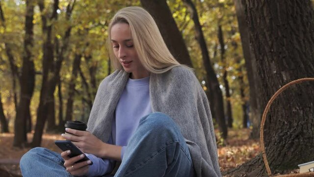 Blonde woman drinks coffee and uses phone in the park at fall on a sunny day