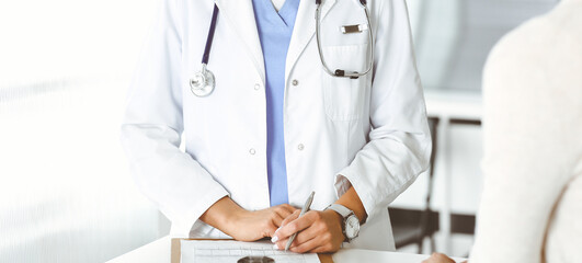 Unknown woman-doctor sitting and communicating with female patient. Physician checks medical history record and exam results, close-up. Healthcare concept