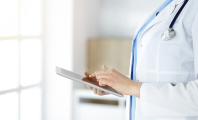 Unknown woman-doctor standing in sunny clinic and using tablet pc, close-up. Data and best service in medicine