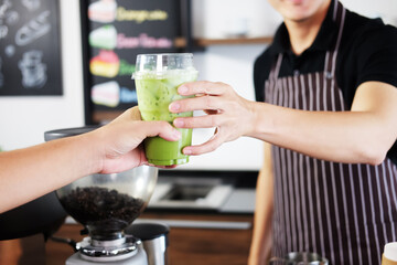 Smiling Asian barista young man serving a plastic glass of iced Matcha green tea and take away to home for customers at counter bar in coffee shop. Start up for bussiness Concept
