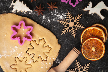 Cookie dough with Christmas cookie cutters, Christmas ornaments. Making gingerbread cookies for winter holidays. Dry orange slices,  candy cones and wooden New Year decoration on the black background.