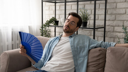 Stressed young man in glasses sitting on sofa, suffering from overheating or hot summer weather without air conditioner, using paper fan cooling himself, breathing fresh air, relaxing alone at home.