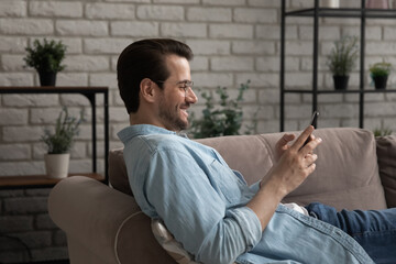 Side view addicted to technology happy young man relaxing on comfortable couch with smartphone in hands, enjoying communicating with friends in social network or using mobile dating application.