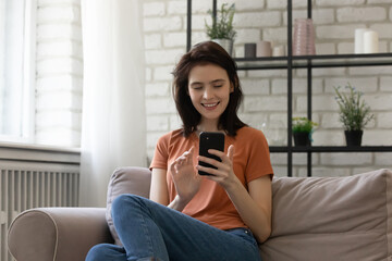 Relaxed millennial smiling woman sitting on sofa, spending free leisure time communicating in social networks with friends, scrolling news feed in mobile phone app, technology addiction concept.