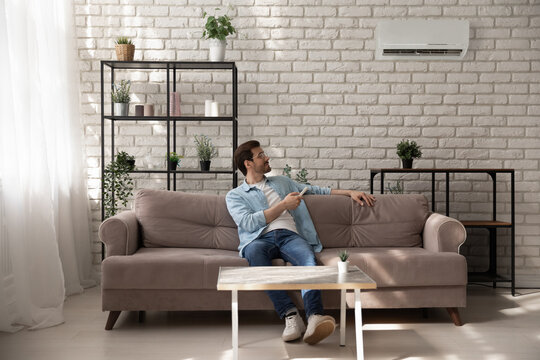 Full length relaxed young man sitting on comfortable sofa, turning on climate control system with remote controller, feeling satisfied with fresh conditioned air during hot summer time weather at home