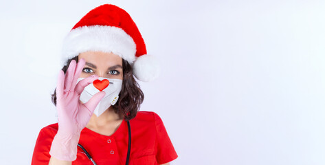 Close-up portrait of a female doctor in a medical protective mask, Santa hat and pink nitrile gloves, as well as showing a heart, a symbol of love, Healthy heart, cardiology. Isolate. Copy space
