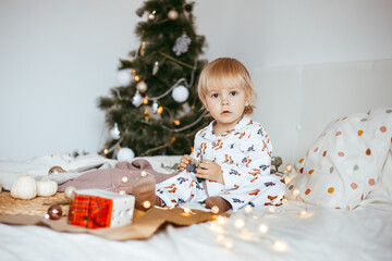 the child's first Christmas. a pensive little boy is lying in his holiday pyjamas on a bed against the background of a brightly decorated fir tree with a Golden light garland.