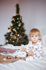 the child's first Christmas. a pensive little boy is lying in his holiday pyjamas on a bed against the background of a brightly decorated fir tree with a Golden light garland.