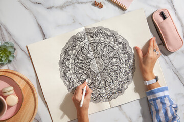 Top view of colored mandala art book with colorful markers, pen, pink pen case and a cup of coffee and macaroons. White marble background.