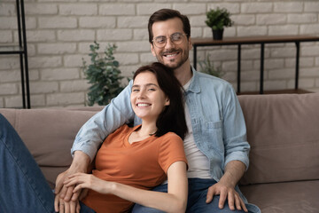 Portrait of smiling young caucasian loving family couple renters homeowners relaxing on comfortable sofa in living room, enjoying spending free weekend time together at home, looking at camera.