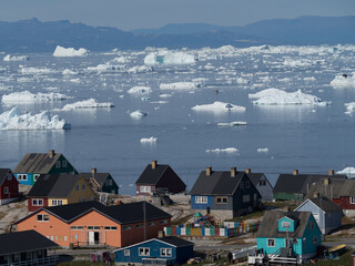 View across a Greenland bay shows drifting ice bergs and traditional coloured housing in the foreground. © Tony Skerl