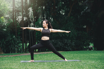 Asian woman practicing yoga in Warrior Pose (Virabhadrasana) on the mat in outdoor park.