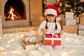 Obraz na płótnie Canvas Little girl opening christmas present, keeping fingers on ribbons, looking at gift box with smile, child wearing santa claus hat posing on floor near fireplace and xmas tree.