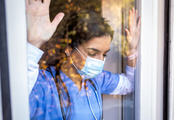 Portrait of exhausted nurse with mask standing behind the glass.