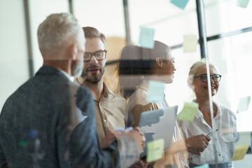 Group of multiracial coworkers standing in the modern office and putting colorful sticky notes on a glass wall and discussing project, working together and sharing ideas