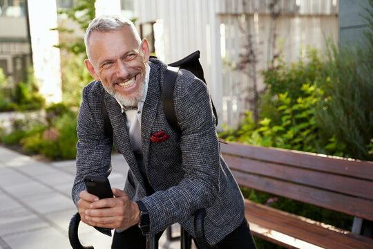 Happy Middle Aged Businessman In Stylish Suit Smiling While Holding His Smartphone, Standing With Bicycle Outdoors In The Daytime