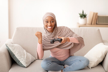 Weekend pastimes. Happy black woman in hijab with joystick playing videogames indoors, making YES gesture © Prostock-studio