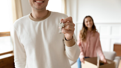 Close up husband holding keys, wife on background with cardboard box. Young family buying first house together, new first property purchase, bank lending, ownership, life changes, beginnings concept